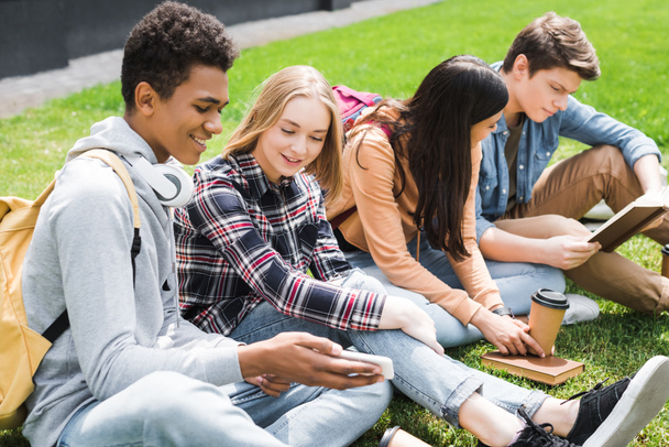 Lächelnde und glückliche Teenager, die auf Gras sitzen, aufs Smartphone schauen und Bücher lesen  - Foto, Bild