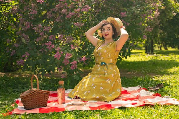 симпатичная plus size девушка в симпатичном винтажном желтом платье и наслаждающаяся отдыхом в летнем парке на зеленой траве. Кудрявая пинающая женщина в ретро-платье отдыхает на природе рядом с лилаком
 - Фото, изображение