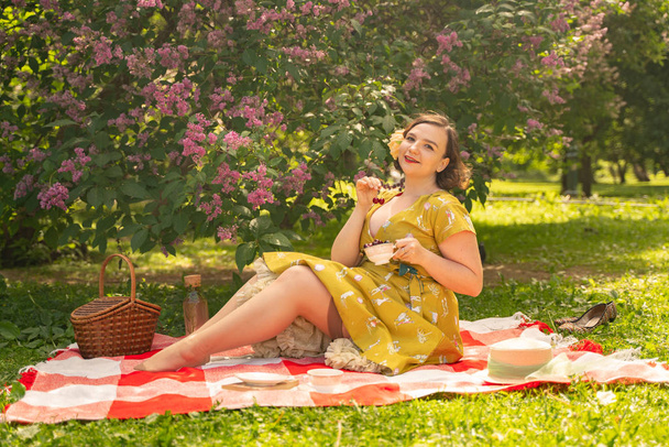 милий плюс розмір біла дівчина в милій вінтажній жовтій сукні і насолоджуватися відпочинком в літньому парку на зеленій траві. пишна піжамна жінка в ретро-сукні відпочиває на природі біля бузку
 - Фото, зображення