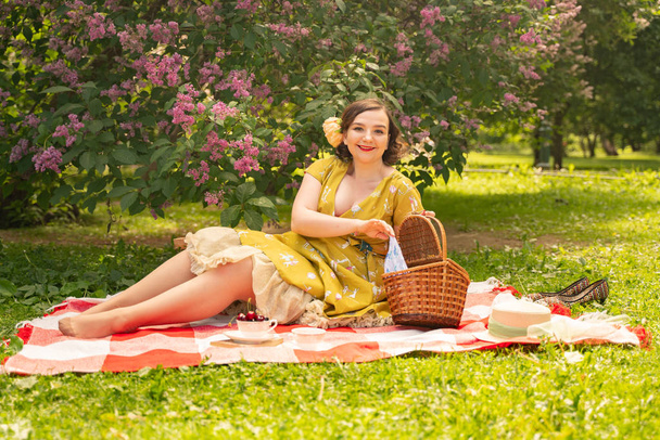 cute plus size caucasian girl wearing a cute vintage yellow dress and enjoying a vacation in the summer Park on the green grass. curvy pinup woman in retro dress has rest on the nature near the lilac - Zdjęcie, obraz