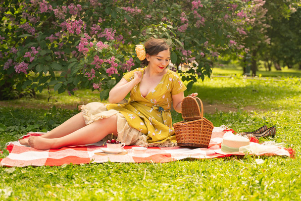 cute plus rozmiar kaukaski dziewczyna sobie cute rocznika żółty strój i korzystających z wakacji w letnim parku na zielonej trawie. Krzywego Pinup Kobieta w retro sukienka ma odpocząć na naturze w pobliżu bzu - Zdjęcie, obraz