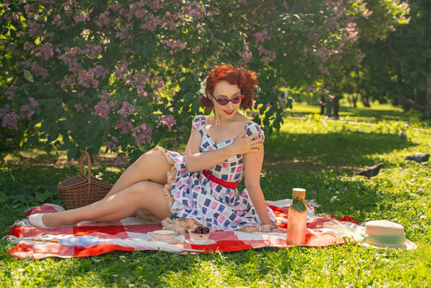 zrzavý šťastný špendlík v letních šatech a klasické punčochy s šekem vzadu sedí na přikrývce na zelené trávě v městském parku vedle šeříku - Fotografie, Obrázek