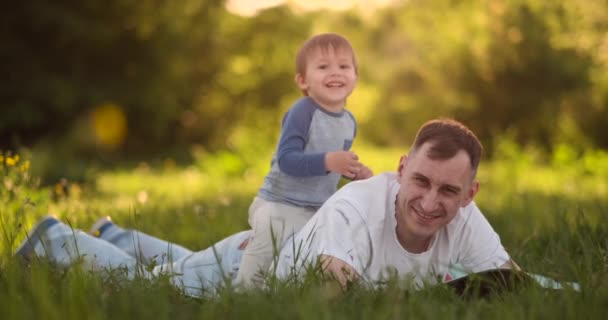 Amoroso jovem pai e filho brincam na grama no verão ao pôr do sol em câmera lenta
 - Filmagem, Vídeo