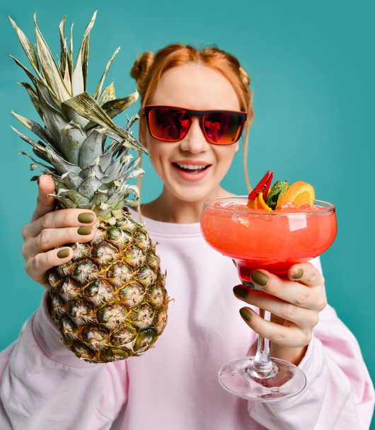 Cheerful blonde woman in modern red sunglasses handed us demonstrates a strawberry margarita cocktail and a big pineapple - Foto, Imagem