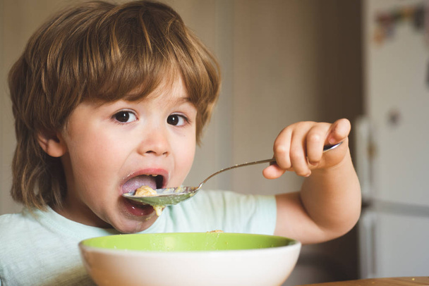 Πεινασμένο αγοράκι που τρώει. Το χαρούμενο μωρό τρώει το ίδιο το φαγητό με κουτάλι. Νόστιμο παιδικό πρωινό. Το μωρό τρώει φαγητό στην κουζίνα. Το χαρούμενο αγοράκι τρώει μόνο του υγιεινά φαγητά.. - Φωτογραφία, εικόνα