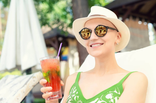Young happy woman holding fresh cold strawberry mojito cocktail enjoying vacation at resort on hot sunny day. young caucasian bald woman in hat and swimsuite enjoy life after surviving breast cancer - Valokuva, kuva