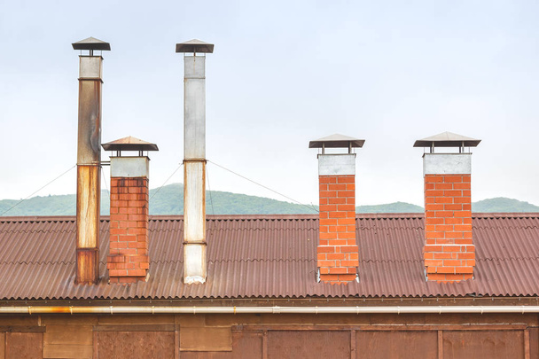 Brick chimneys and tin ventilation pipes on the roof of a wooden hut on a background of green hills on a cloudy day - Photo, Image