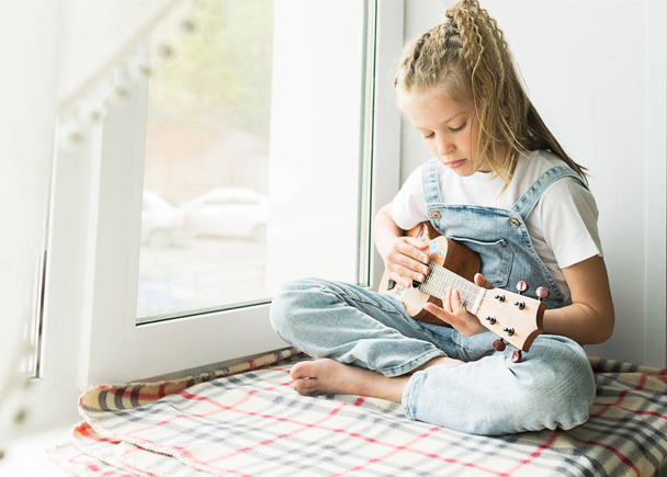 Mała dziewczynka siedzi przy oknie w domu grając na gitarze. Selektywne skupienie. Koncepcja muzyki i sztuki. - Zdjęcie, obraz