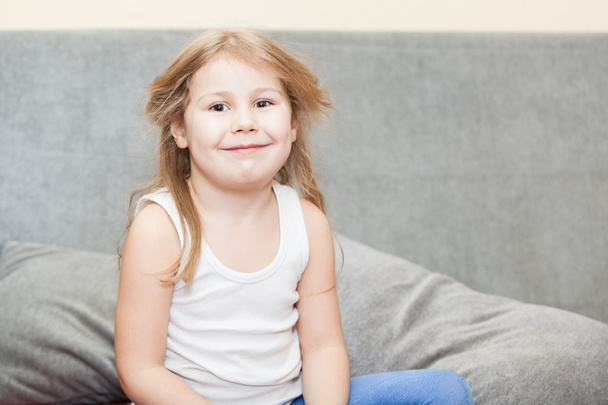 Little Caucasian girl with disheveled hair sitting on the couch with a sly grin - Foto, Imagem