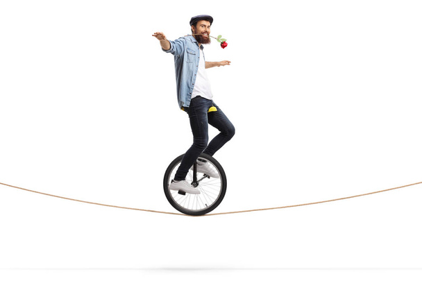 Full length shot of a bearded young man riding a unicycle on a rope and holding a red rose with his mouth isolated on white background - Valokuva, kuva