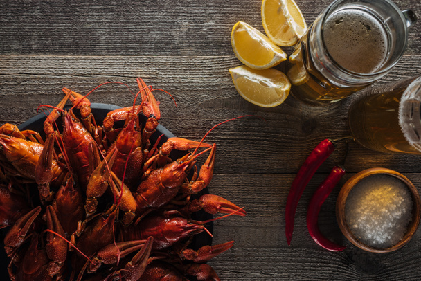 top view of red lobsters, lemon slices, peppers, glasses with beer and salt on wooden surface - Photo, Image