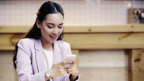 smiling and pretty asian businesswoman in suit using smartphone in cafe  - Video