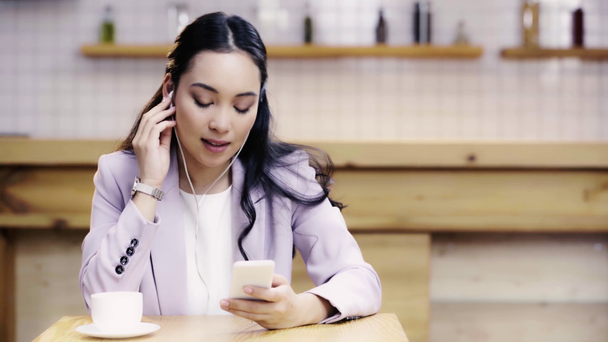 hymyilevä ja kaunis aasialainen nainen puku kuuntelee musiikkia ja käyttää älypuhelinta kahvilassa
  - Materiaali, video