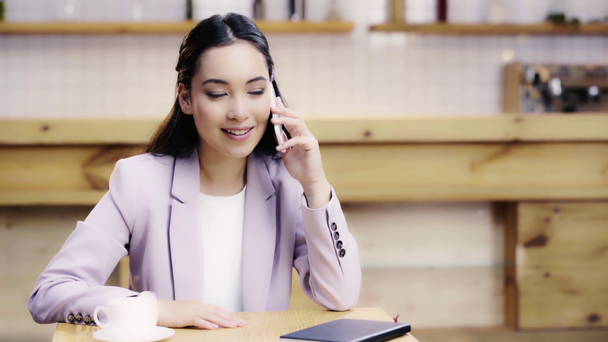 χαμογελαστή και όμορφη Ασιατική γυναίκα στο κοστούμι μιλάει στο smartphone στο καφέ  - Πλάνα, βίντεο