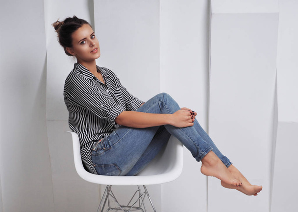 portrait of beautiful young woman in striped shirt sitting on chair in front of white wall - Photo, Image