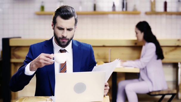 серьезный бизнесмен смотрит на документы и экран ноутбука, пьет кофе, деловая женщина печатает на компьютере на заднем плане
  - Кадры, видео