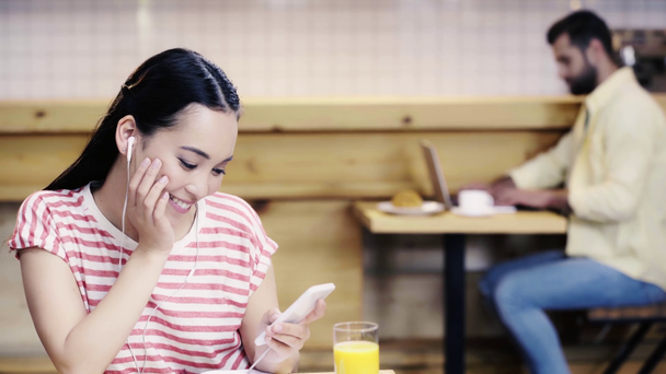 glimlachend en mooie Aziatische vrouw in t-shirt luisteren muziek en man met behulp van smartphone en laptop op de achtergrond  - Video