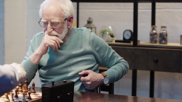 homem idoso barbudo pensivo em óculos jogando xadrez com amigo e pressionando o botão no relógio de xadrez na mesa
 - Filmagem, Vídeo