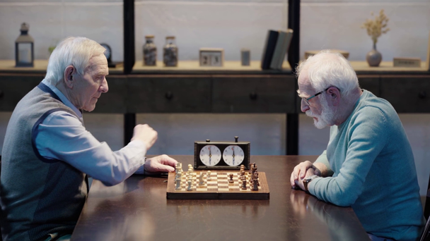 side view of two pensive senior men sitting at table, playing chess and pressing button on chess clock in living room - Filmati, video