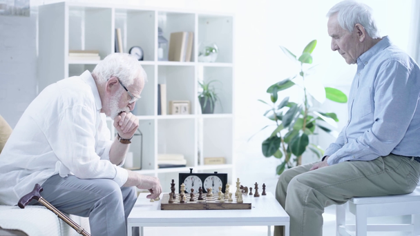vista lateral de dois homens idosos pensativos jogando xadrez na mesa na sala de estar
 - Filmagem, Vídeo