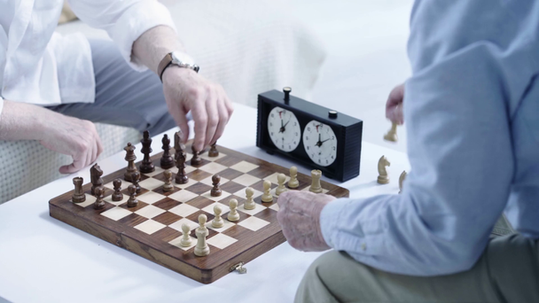 vista parcial de dos hombres mayores en camisas poniendo figuras en el tablero de ajedrez en la mesa en la sala de estar
 - Metraje, vídeo