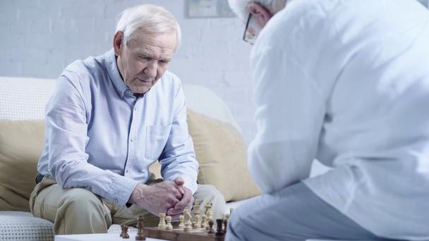two senior men in shirts talking while playing chess in living room - Imágenes, Vídeo