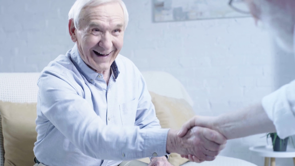 hombre mayor riendo en camisa sentado en el sofá y estrechando la mano con un amigo en la sala de estar
 - Metraje, vídeo