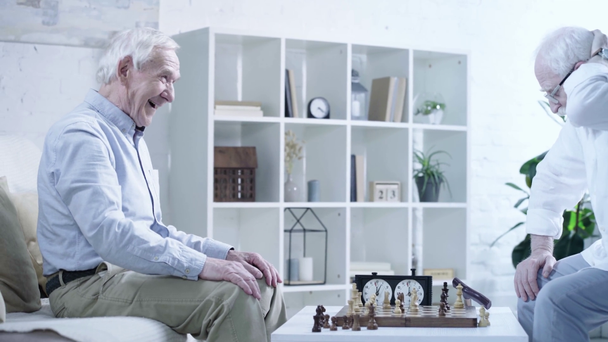 side view of senior man laughing while playing chess with pensive friend in living room - Footage, Video