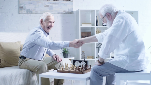 szczęśliwy starszy mężczyzna siedzi w pobliżu szachu, potrząsając rękami z przeciwnikiem i śmiech w salonie - Materiał filmowy, wideo