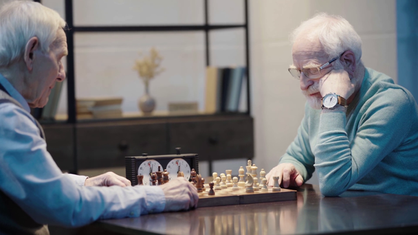 pensive bearded senior man in glasses playing chess and pressing button on chess clock - Filmati, video