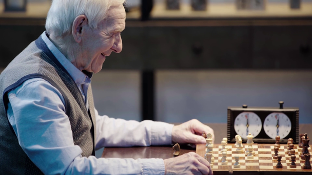 Seitenansicht eines älteren Mannes in Weste, der mit einem Freund Schach spielt, Knopf an der Schachuhr drückt und im Wohnzimmer lacht - Filmmaterial, Video