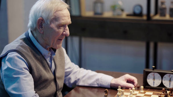 cheerful senior man in vest playing chess with opponent and smiling in living room - Footage, Video