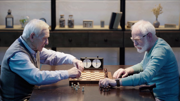 vista lateral de dois homens seniores colocando figuras no tabuleiro de xadrez na mesa de madeira
 - Filmagem, Vídeo