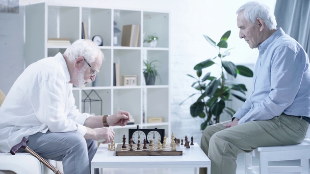Seitenansicht zweier älterer Männer, die Schach spielen und Knopf an der Schachuhr im Wohnzimmer drücken - Filmmaterial, Video