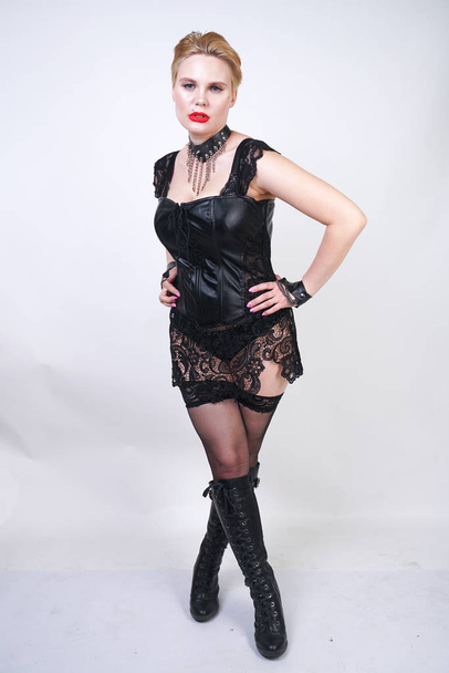 hot fashion young plus size woman wearing leather dress with black lace and sexy stockings and posing on white studio background alone. short hair curvy adult girl standing in mistress bdsm costume. - Photo, Image