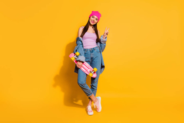 Full length body size photo beautiful she her lady amazing nice look hand arm skate board dangerous sport v-sign symbol wear casual jeans denim jacket shoes pink hat isolated yellow vivid background - Photo, Image