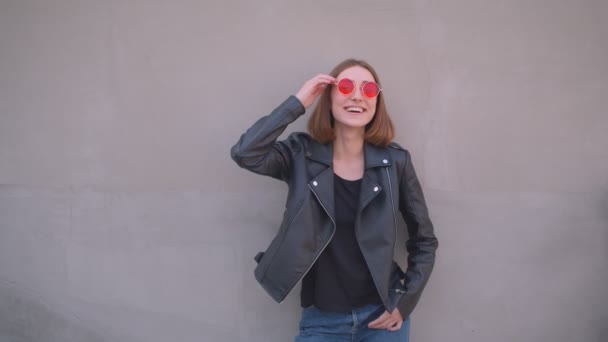 Zbliżenie portret młodego cute kaukaski dziewczyna w skórzanej kurtce i czerwone okulary słoneczne uśmiechnięty i śmiech stwarzające przed kamerą na zewnątrz w miejskim mieście - Materiał filmowy, wideo