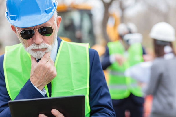Serious, worried, senior gray haired engineer or businessman working on a tablet on construction site - Фото, изображение