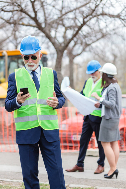 Serieuze Senior ingenieur of zakenman die zijn smartphone gebruikt tijdens het inspecteren van een bouwplaats - Foto, afbeelding
