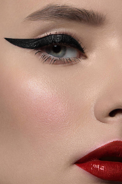 Close-up beauty of a half female face with creative fashion evening make-up. Black arrows on the eyes and extremely long eyelashes, on plump lips matte scarlet lip color. Well-groomed skin after spa - Foto, imagen