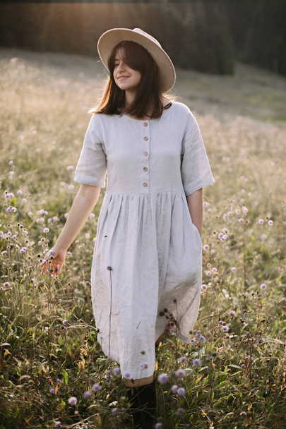 Chica con estilo en vestido rústico y sombrero caminando entre flores silvestres i
 - Foto, imagen