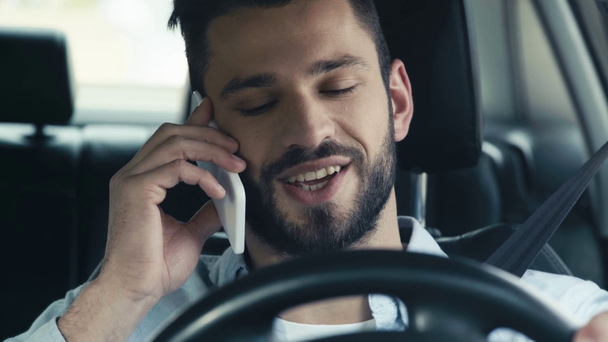 selektivní zaměření pohledného muže, který mluví na telefonu, s úsměvem a přidržením volantu  - Záběry, video