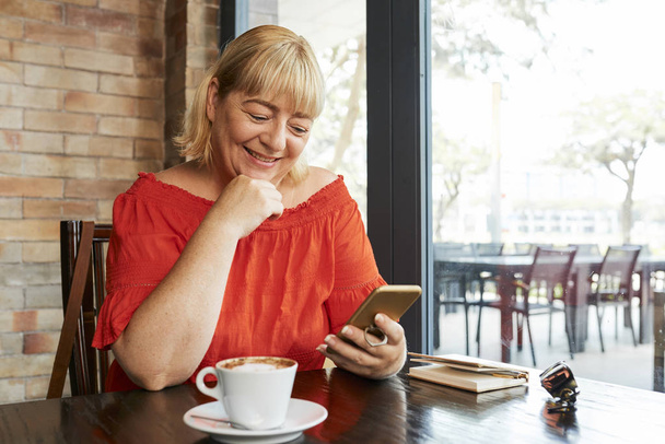 Щаслива зріла жінка відпочиває за столом кафе з чашкою кави і читає повідомлення на своєму телефоні
 - Фото, зображення