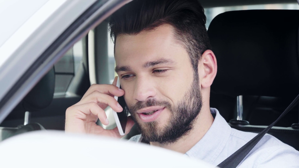 επιλεκτική εστίαση του όμορφου άντρα κάθεται στο αυτοκίνητο, χαμογελώντας και μιλώντας στο smartphone  - Πλάνα, βίντεο