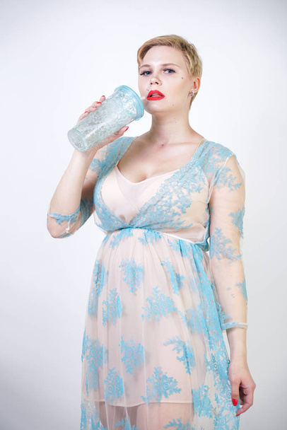 αρκετά χαριτωμένο ξανθιά γυναίκα με κοντά μαλλιά και συν μέγεθος σώμα φορώντας μακρά διαφανή μπλε δαντέλα φόρεμα. κομψό μοντέρνο ενήλικο κορίτσι στο χαριτωμένο νυχτικό στο λευκό φόντο στούντιο μόνο. - Φωτογραφία, εικόνα