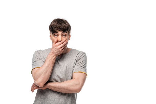 shocked young man looking at camera while covering mouth with hand isolated on white - Photo, Image