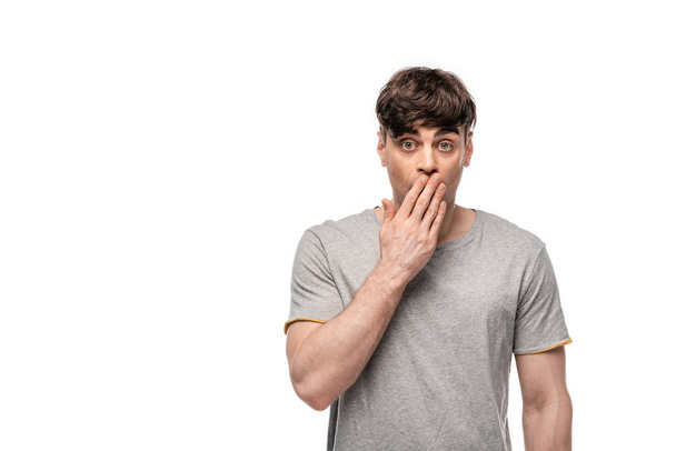 shocked young man looking at camera while covering mouth with hand isolated on white - 写真・画像