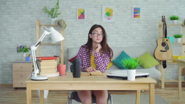 a beautiful Asian girl in glasses with long hair in the living room of a modern house reads a book and uses a voice assistant in preparation for exams - Filmati, video