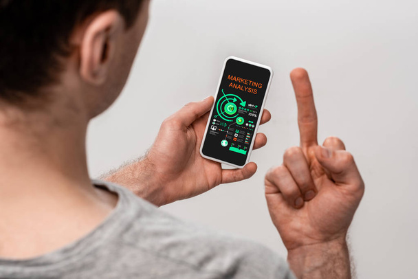 bijgesneden beeld van man met behulp van smartphone met Marketing Analysis app en tonen van middelvinger, geïsoleerd op grijs - Foto, afbeelding
