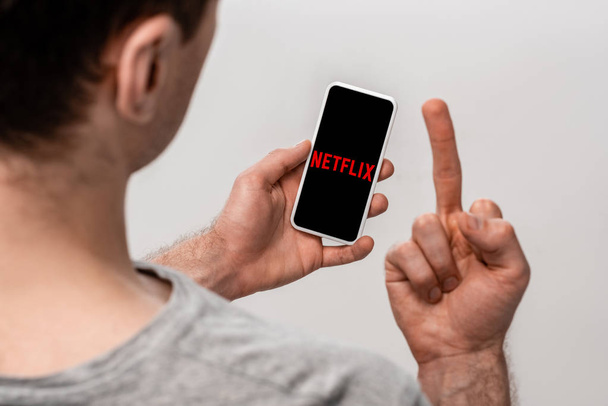 KYIV, UKRAINE - MAY 16, 2019: cropped view of man using smartphone with netflix app and showing middle finger, isolated on grey - Foto, Imagen